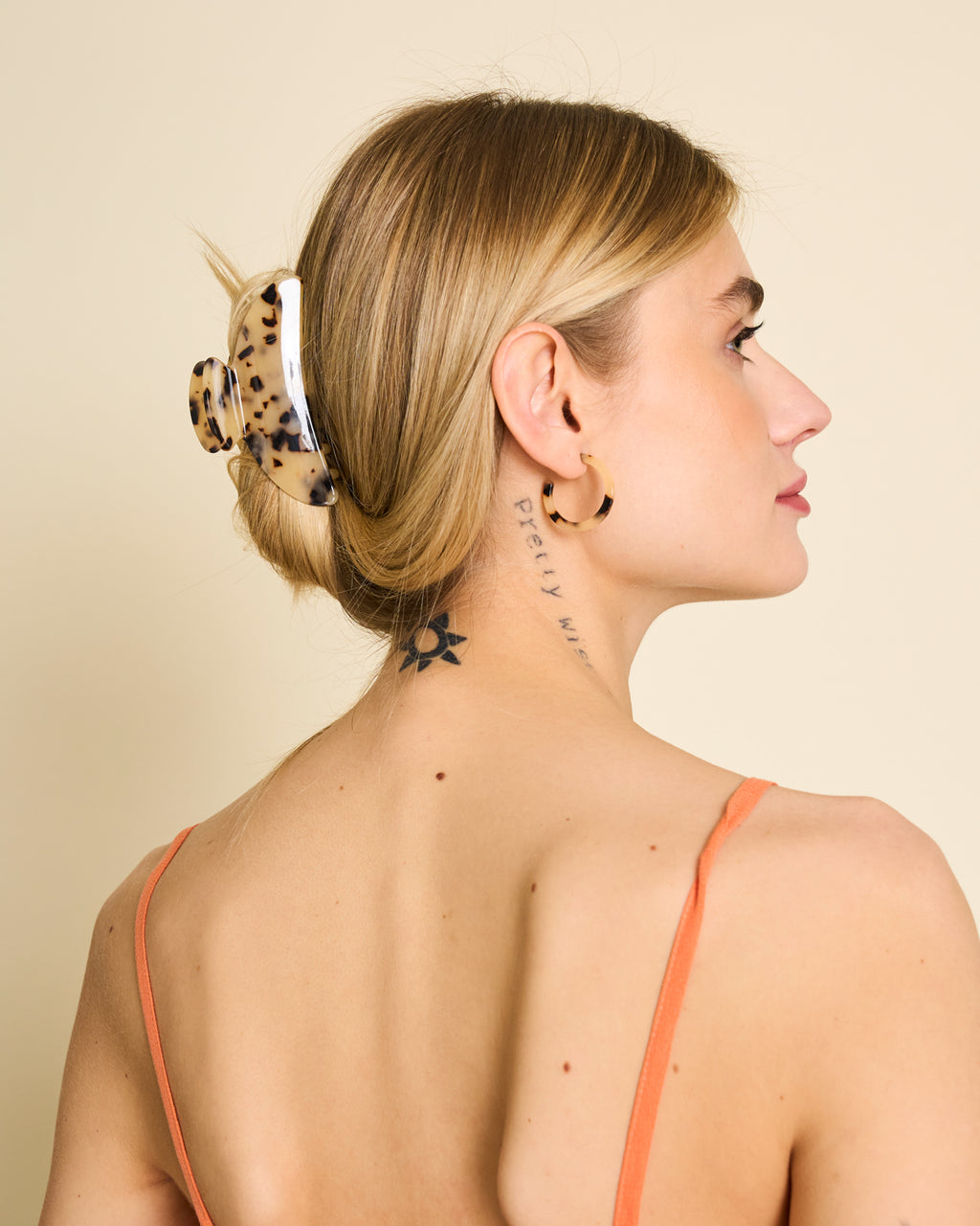 A woman with blonde hair styled in the jan-n-june MIDI HEIRLOOM CLAW BLONDE TORTOISE is seen from behind and in profile, wearing gold hoop earrings, an orange strap top, and showing a star tattoo and script on her neck and back.