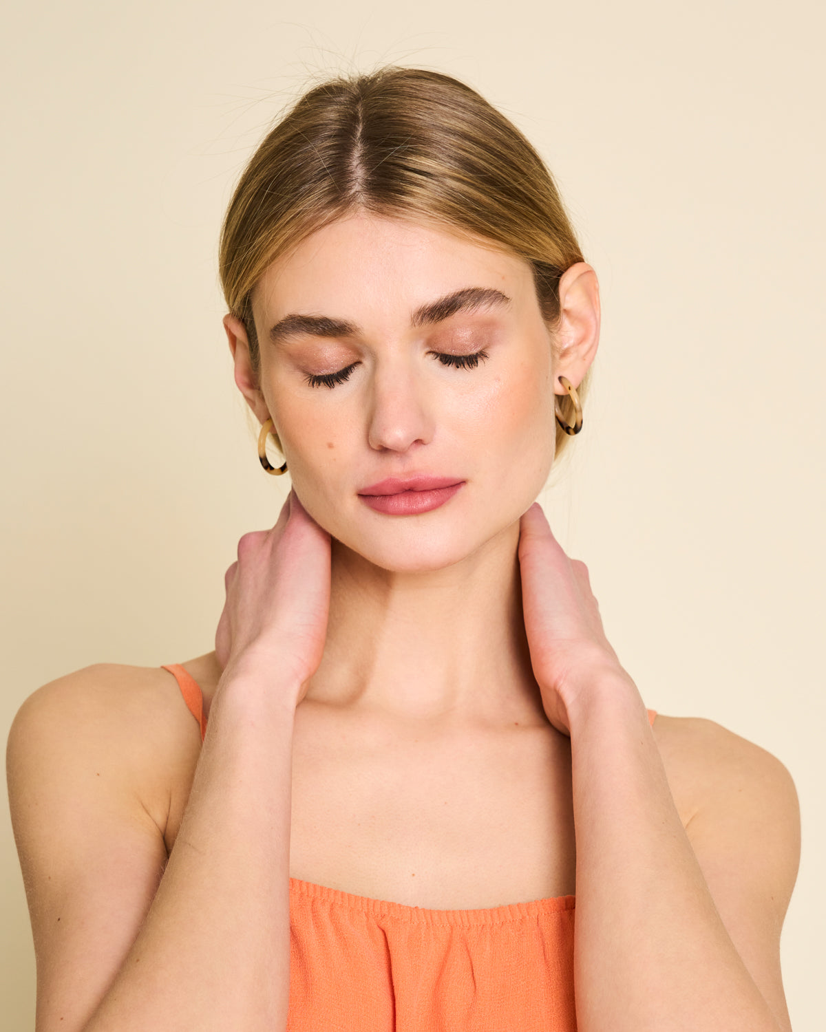 A serene woman with light brown hair wears jan-n-june’s MINI HOOPS BLONDE TORTOISE earrings. She gently holds her neck, dressed in an orange sleeveless top against a beige background.