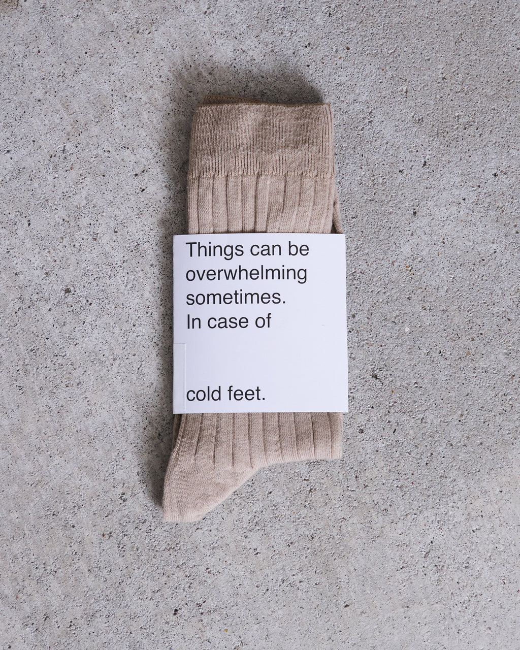 A pair of jan-n-june SOCKS ALMOND in beige organic cotton on a gray concrete surface, topped with a card that says: Things can be overwhelming sometimes. In case of cold feet.