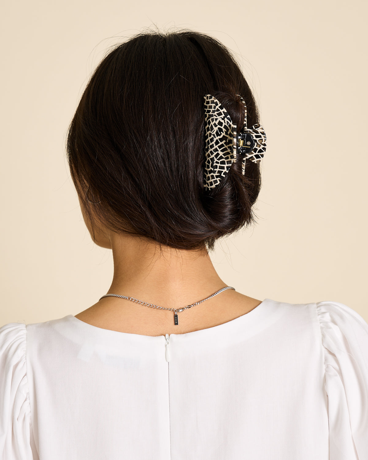 A woman with dark hair in an updo, secured by the jan-n-june MIDI HEIRLOOM CLAW TOKYO CHECKER, is shown from behind. She wears a white puff-sleeve top and a silver necklace.