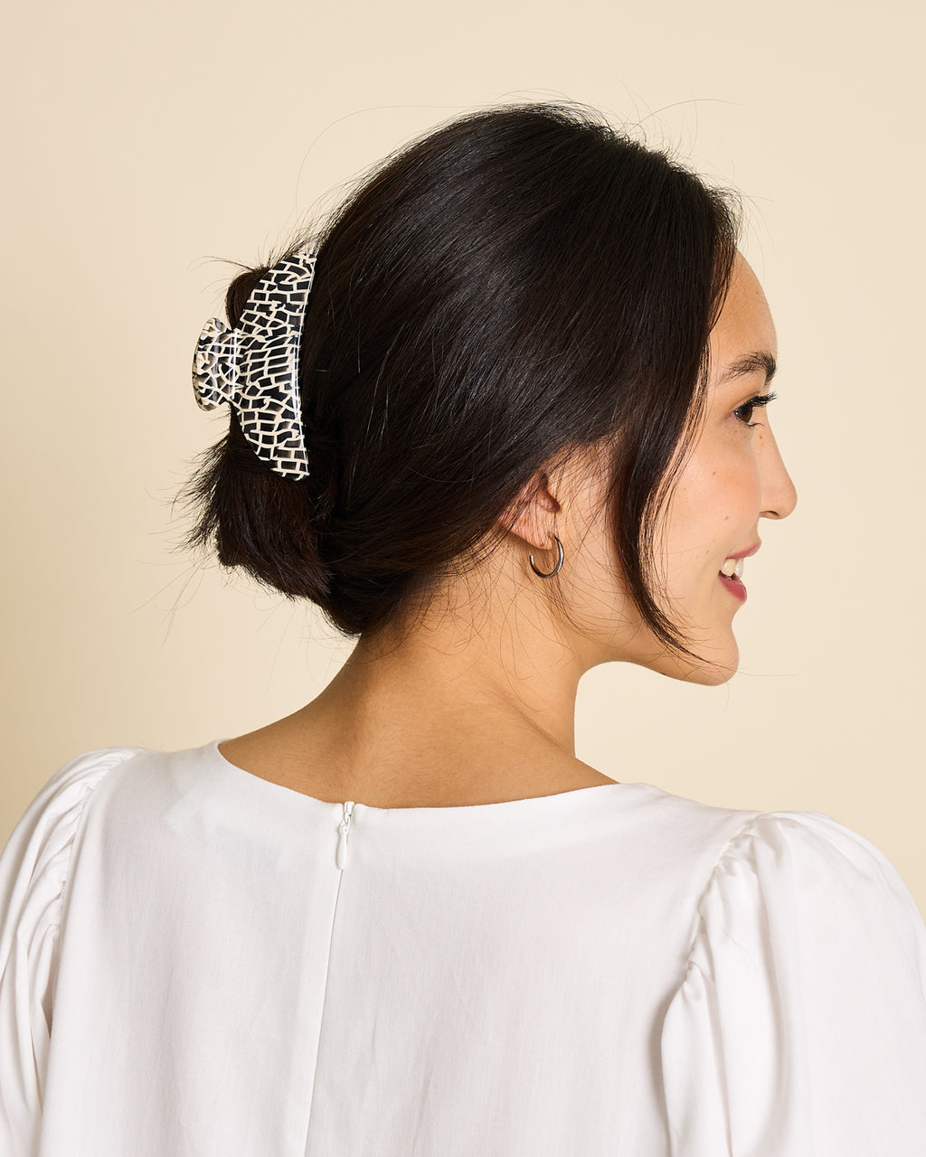 A woman with dark hair in a low updo, secured with the jan-n-june MIDI HEIRLOOM CLAW TOKYO CHECKER, smiles as she looks to the side. She wears a white blouse and gold hoop earring.