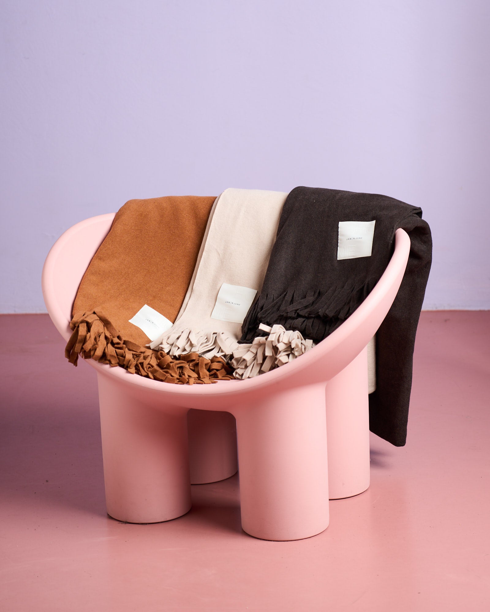 A modern pink chair displays three folded jan-n-june FLEECE BLANKET BRICK organic cotton blankets in camel brown, cream, and dark brown, each with a white label, set against a light purple wall and pink floor.