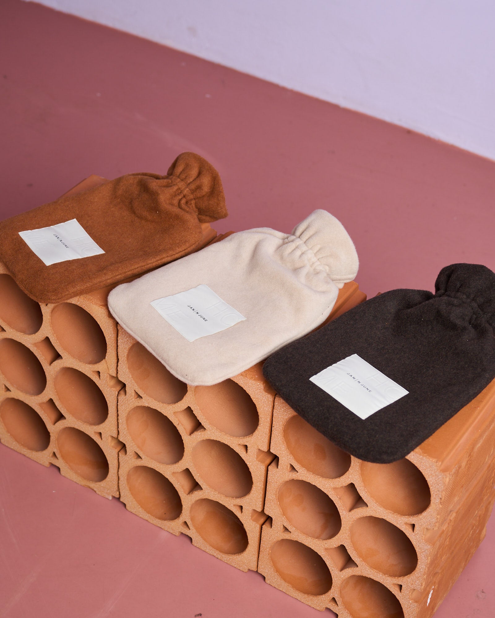 Three jan-n-june HOT WATER BOTTLE COVER MOCCA bottles in brown, cream, and dark gray—each with a sustainable cover—are displayed on stacked hollow bricks against a pink floor and light purple wall background.