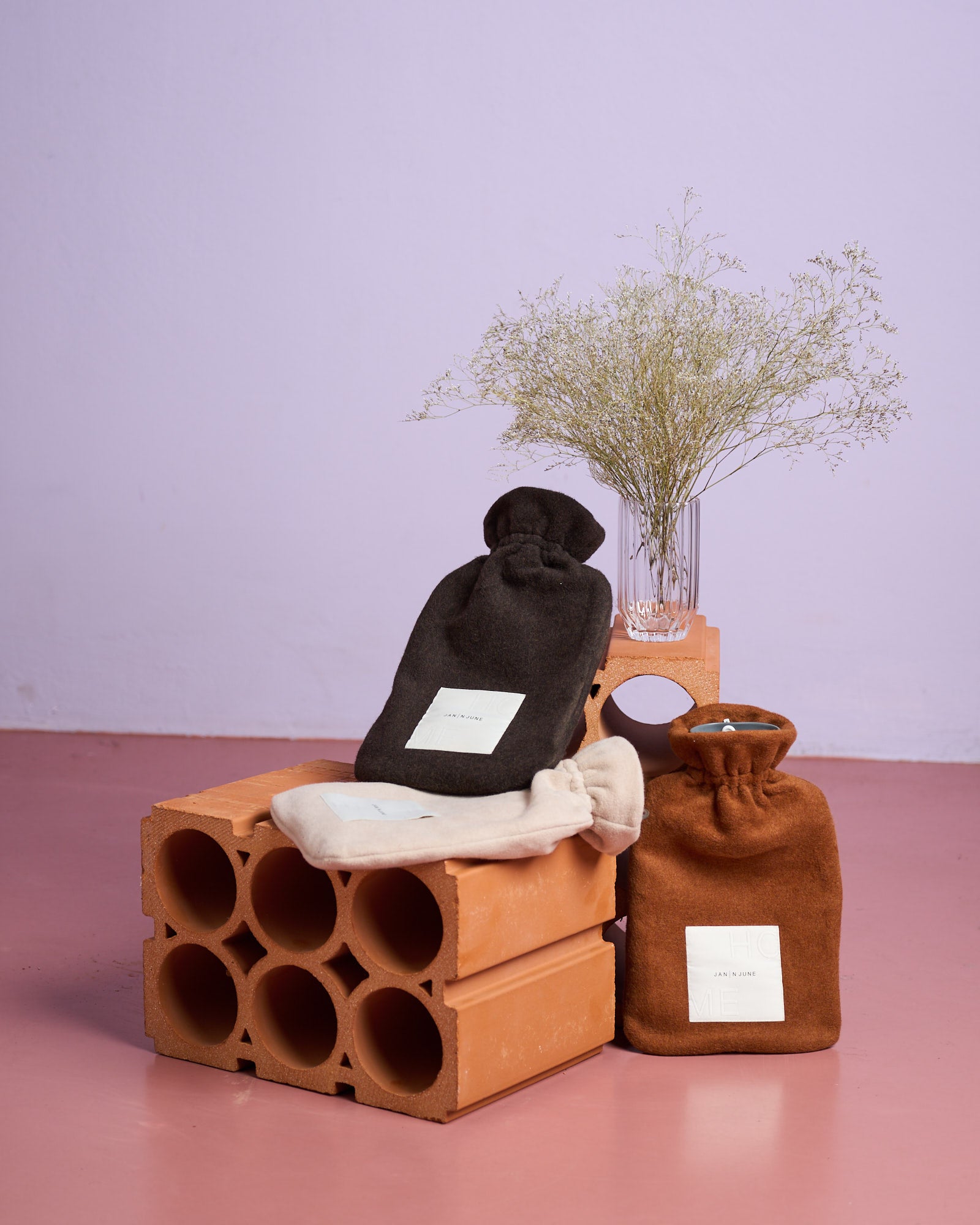 The jan-n-june HOT WATER BOTTLE COVER MOCCA, in organic cotton and rich brown, is displayed on orange concrete blocks beside a clear vase with dried flowers, set against a pale lavender wall and pink floor.