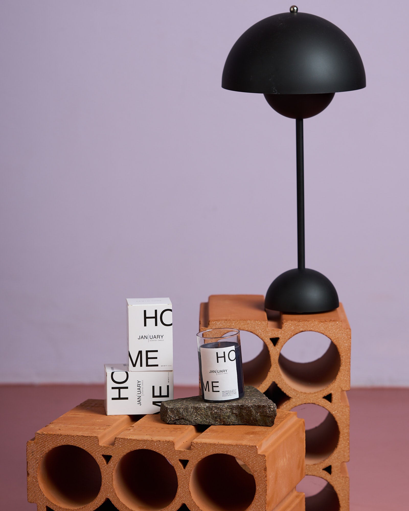 A black modern lamp and jan-n-junes SCENTED CANDLE JAN | UARY—featuring Wild Orange—are displayed on stacked clay bricks with a rock under the candle, set against a light purple and pink background.