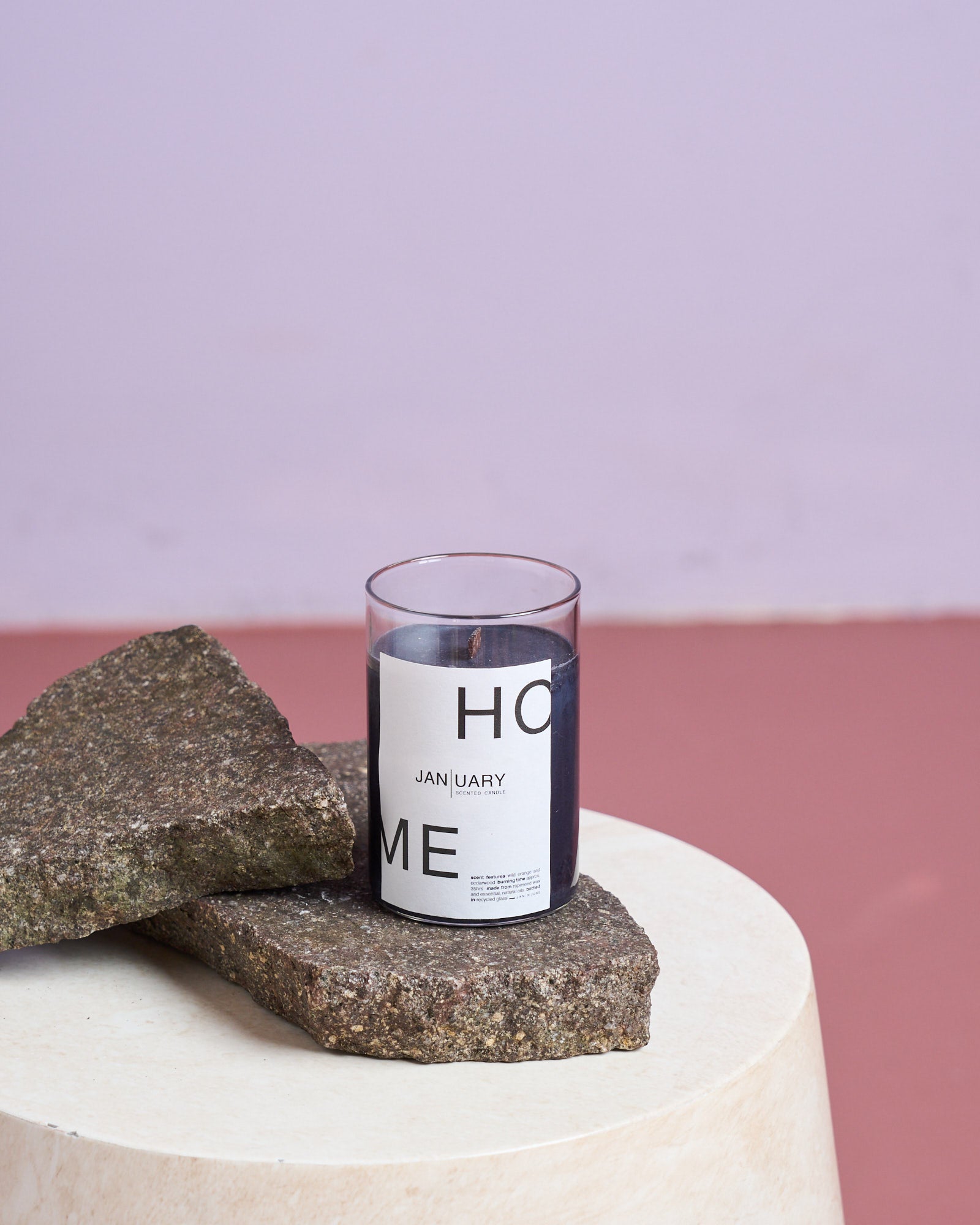 The jan-n-june SCENTED CANDLE JAN | UARY, made with rapeseed wax, rests on two rough stone slabs on a round cream table, set against a mauve wall and reddish floor.