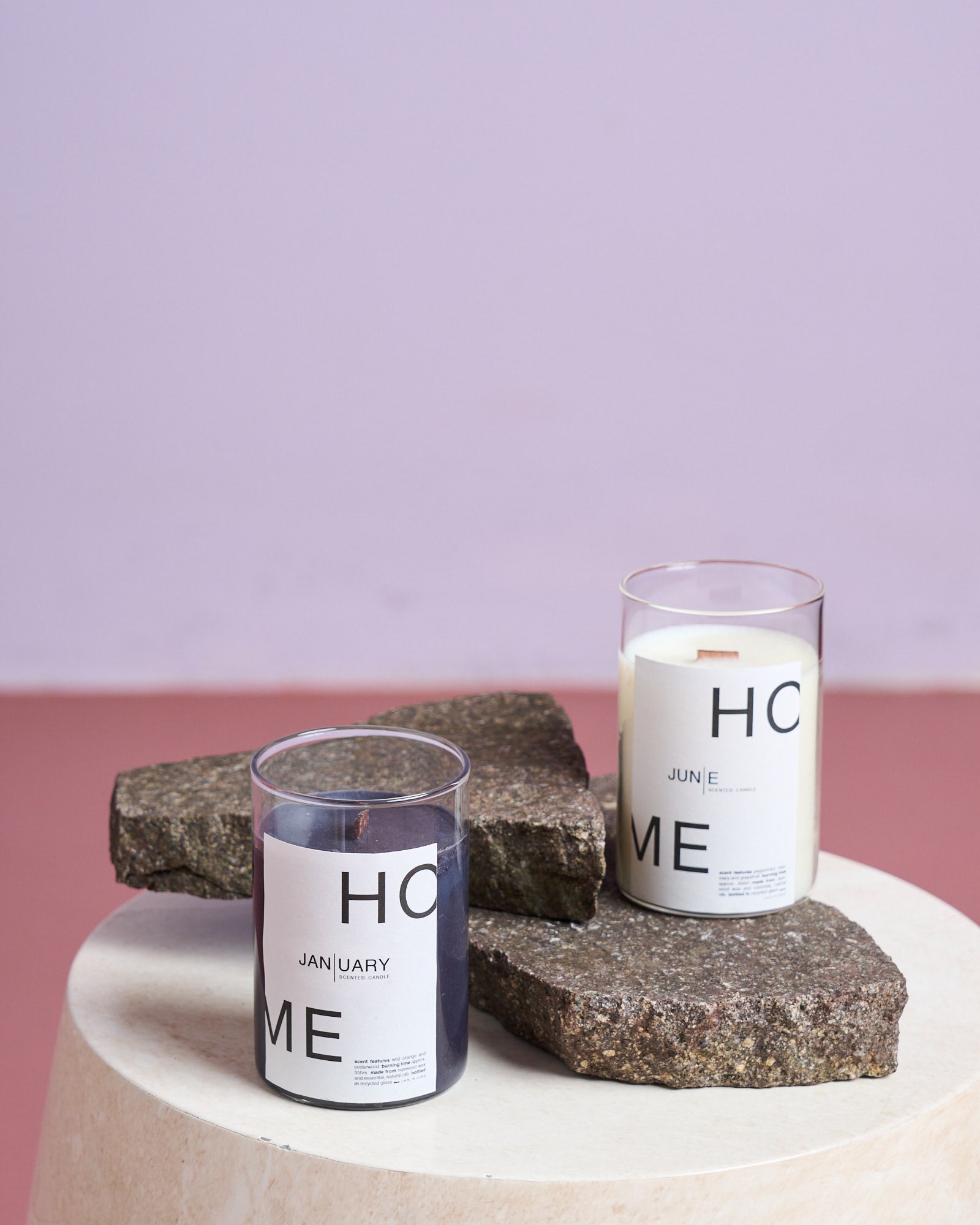 Two SCENTED CANDLE JAN | UARY candles by jan-n-june—one dark, one white, made from rapeseed wax—are displayed on rough stone slabs atop a round white table, with a pastel purple and pink backdrop for a modern look.