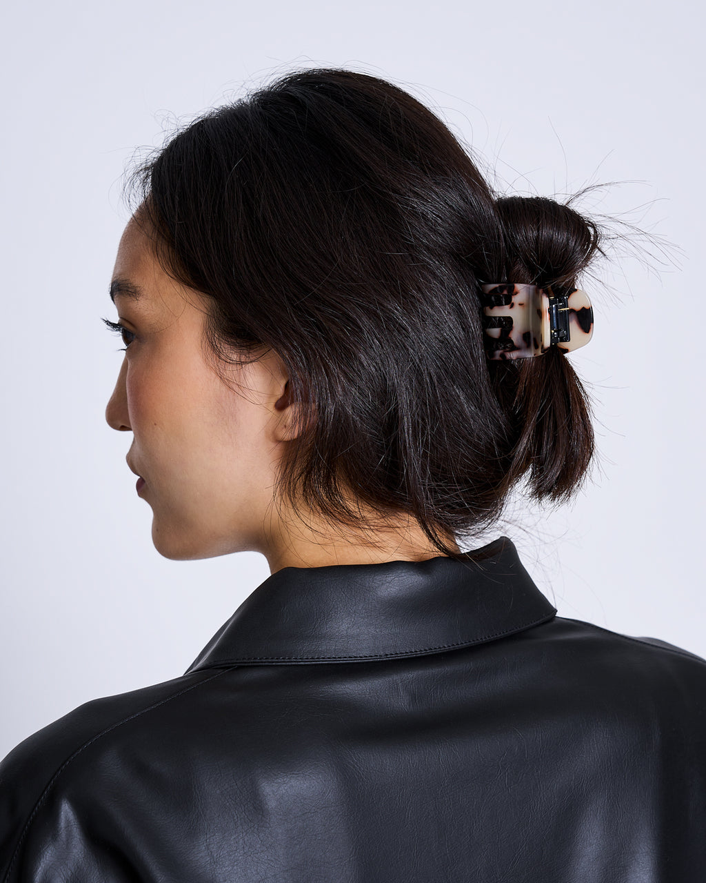 A woman with dark hair in a low bun, secured by the jan-n-june MINI CLAW ASH BLONDE TORTOISE hair accessory, is shown from behind and slightly to the side. She wears a black faux leather jacket against a plain background.
