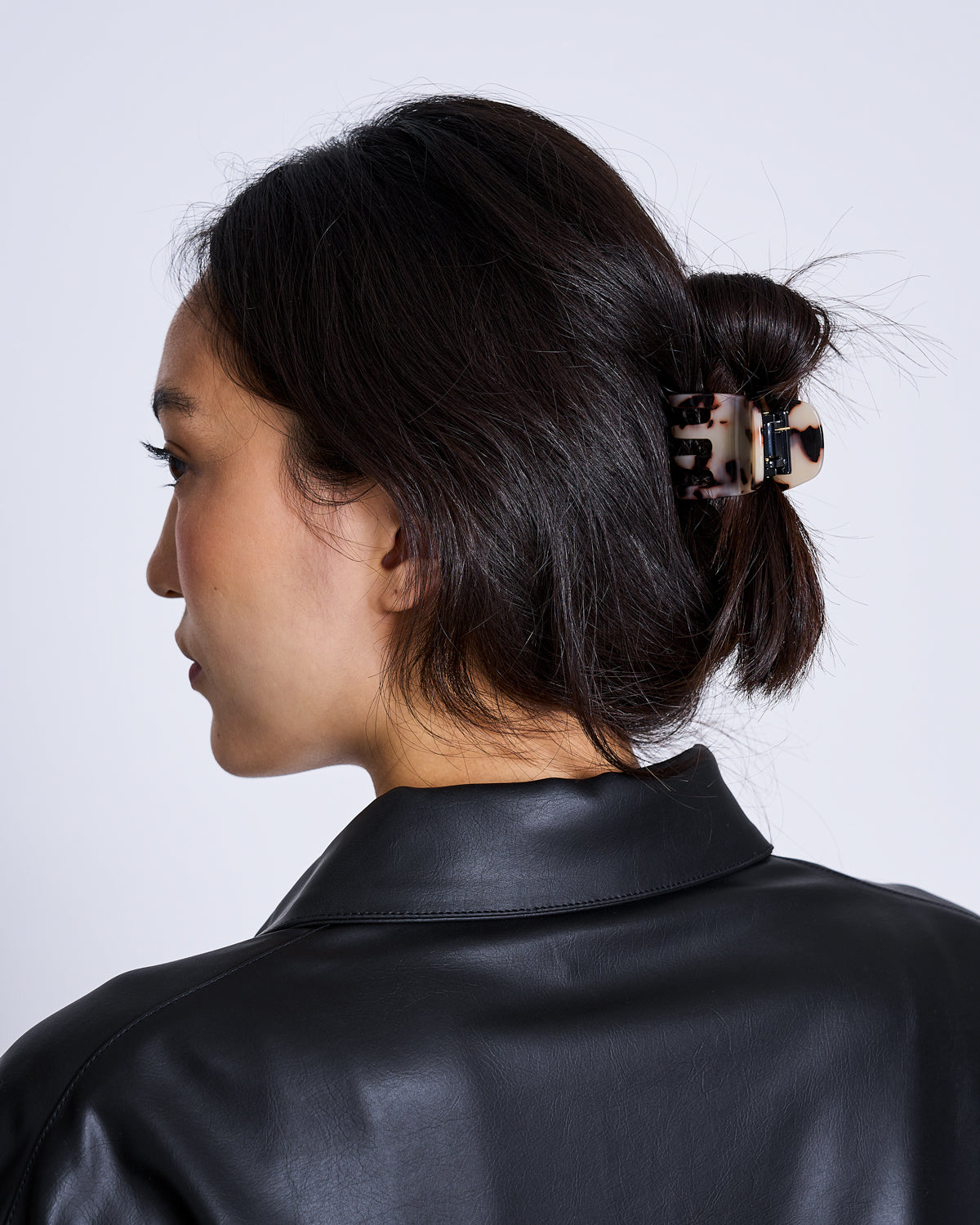 A woman with dark hair in a low bun, secured by the jan-n-june MINI CLAW ASH BLONDE TORTOISE hair accessory, is shown from behind and slightly to the side. She wears a black faux leather jacket against a plain background.