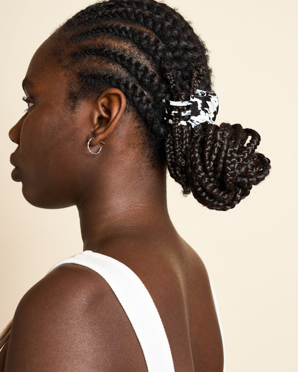 A woman with cornrow braids in a low bun, secured with the jan-n-june MINI CLAW NOIR TORTOISE hair accessory, is shown in profile against a beige background. She wears a white tank top and small hoop earring.