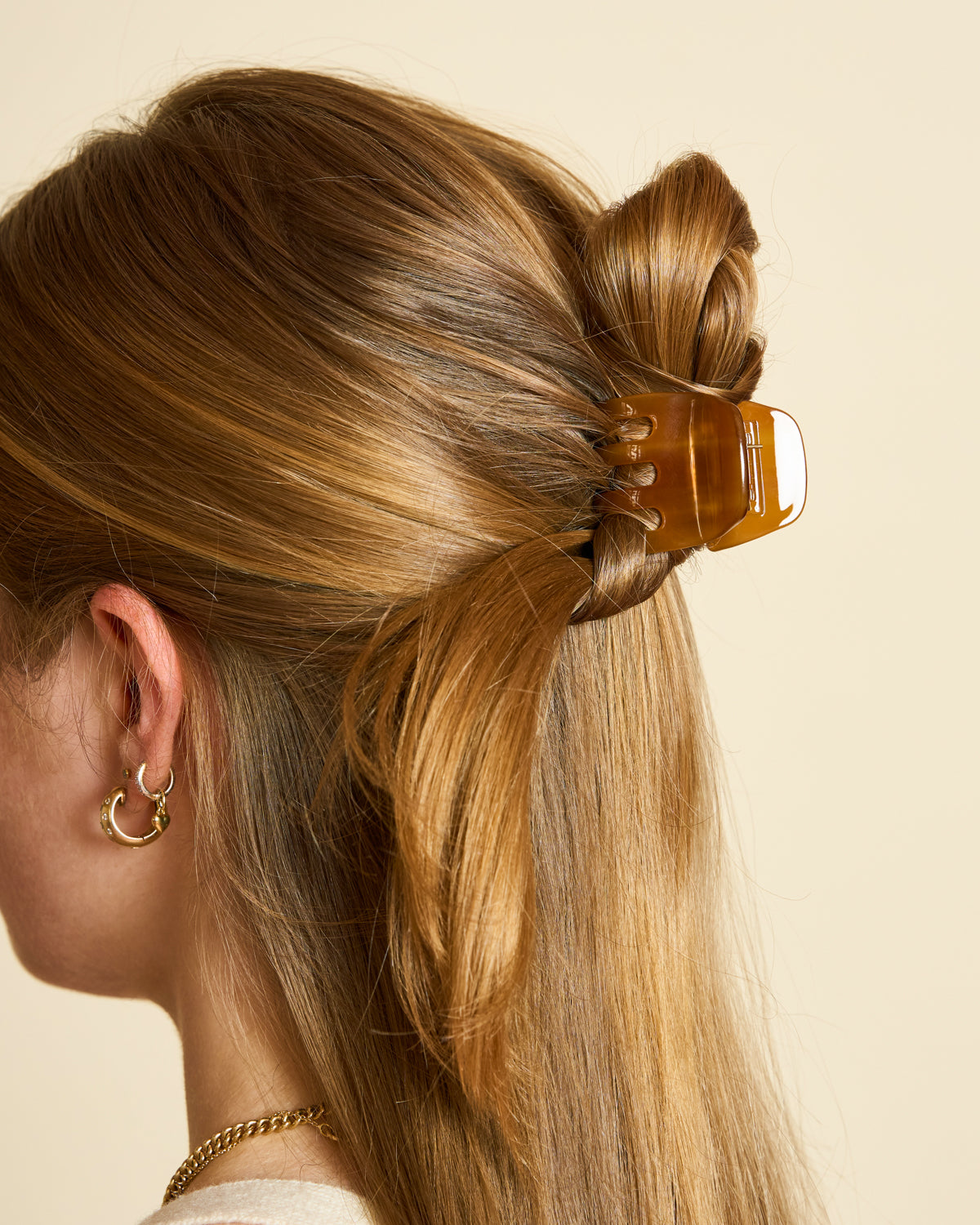 A woman with light brown hair styled in a half-up twist, secured with the jan-n-june MINI CLAW COGNAC, is shown from the back and side against a beige background. She wears gold hoop earrings and a gold chain necklace.