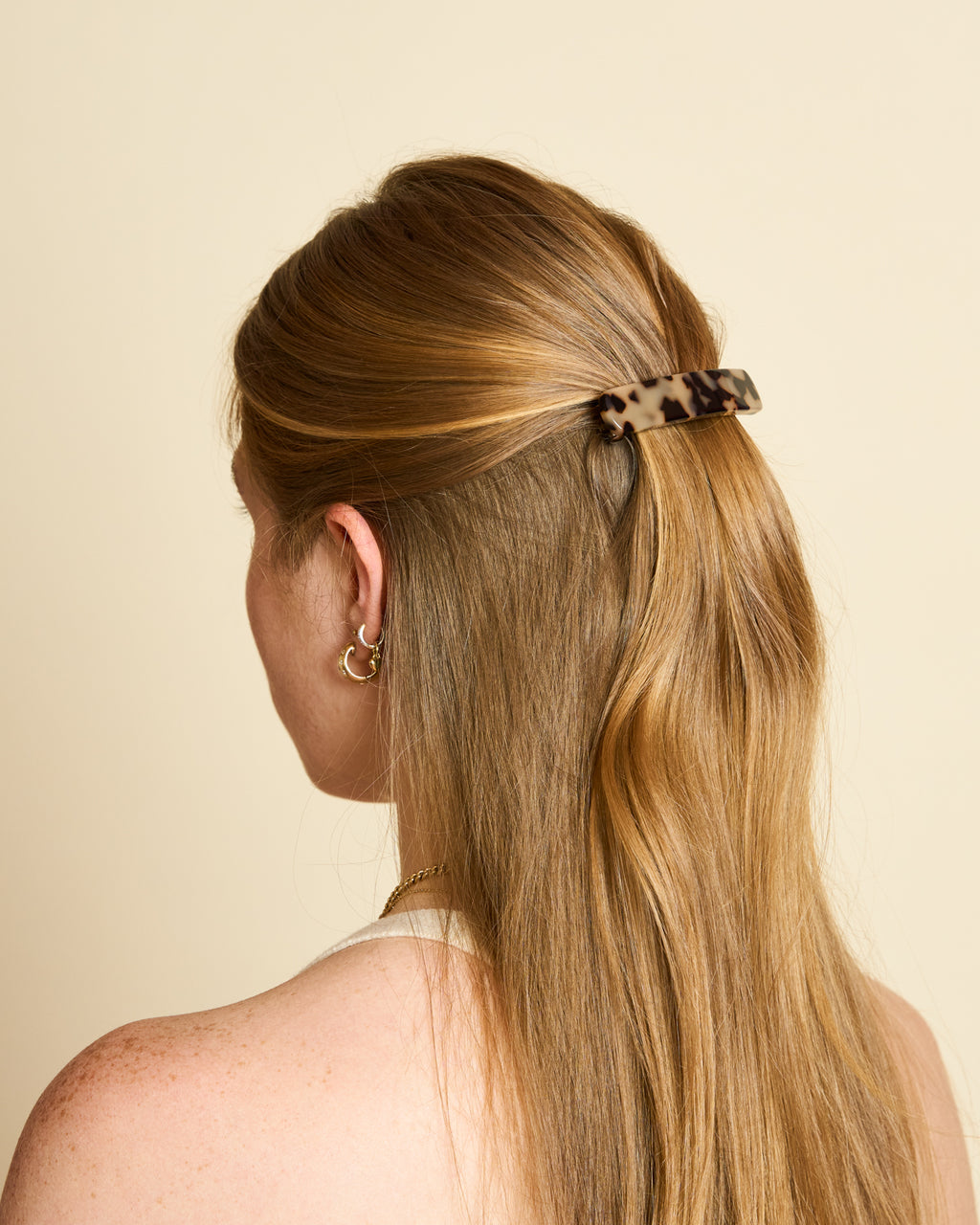 A person with long, light brown hair is seen from behind, their hair partially pulled back with the jan-n-june HEIRLOOM BARRETTE BLONDE TORTOISE, embodying French chic style with gold hoop earrings and a gold necklace.