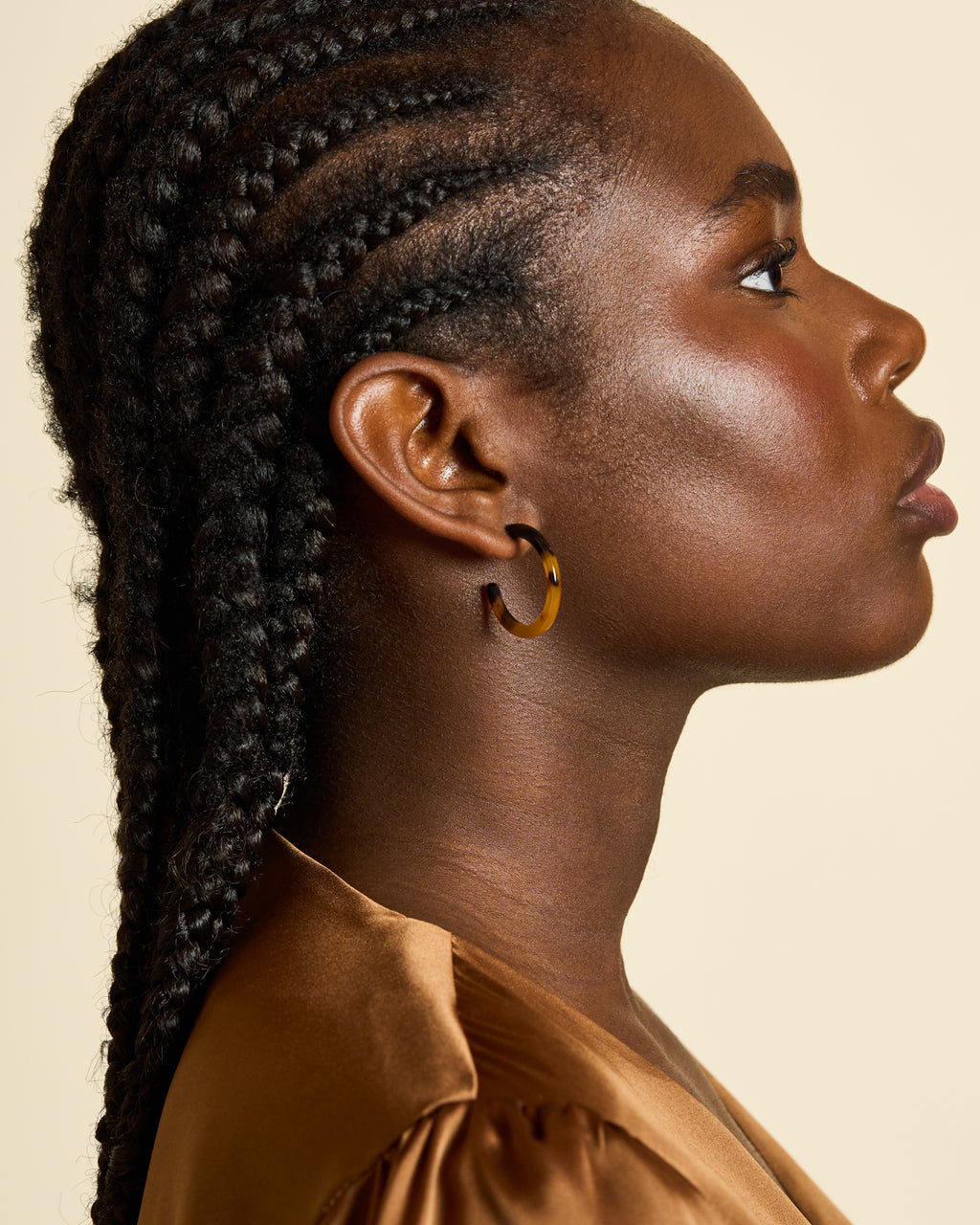 A woman with dark skin and braided hair is shown in profile against a beige background, wearing jan-n-junes MINI HOOPS CLASSIC TORTOISE earrings and a silky brown top. Her expression is calm and focused.