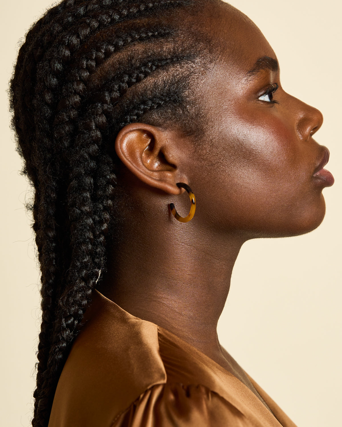 A woman with dark skin and braided hair is shown in profile against a beige background, wearing jan-n-junes MINI HOOPS CLASSIC TORTOISE earrings and a silky brown top. Her expression is calm and focused.