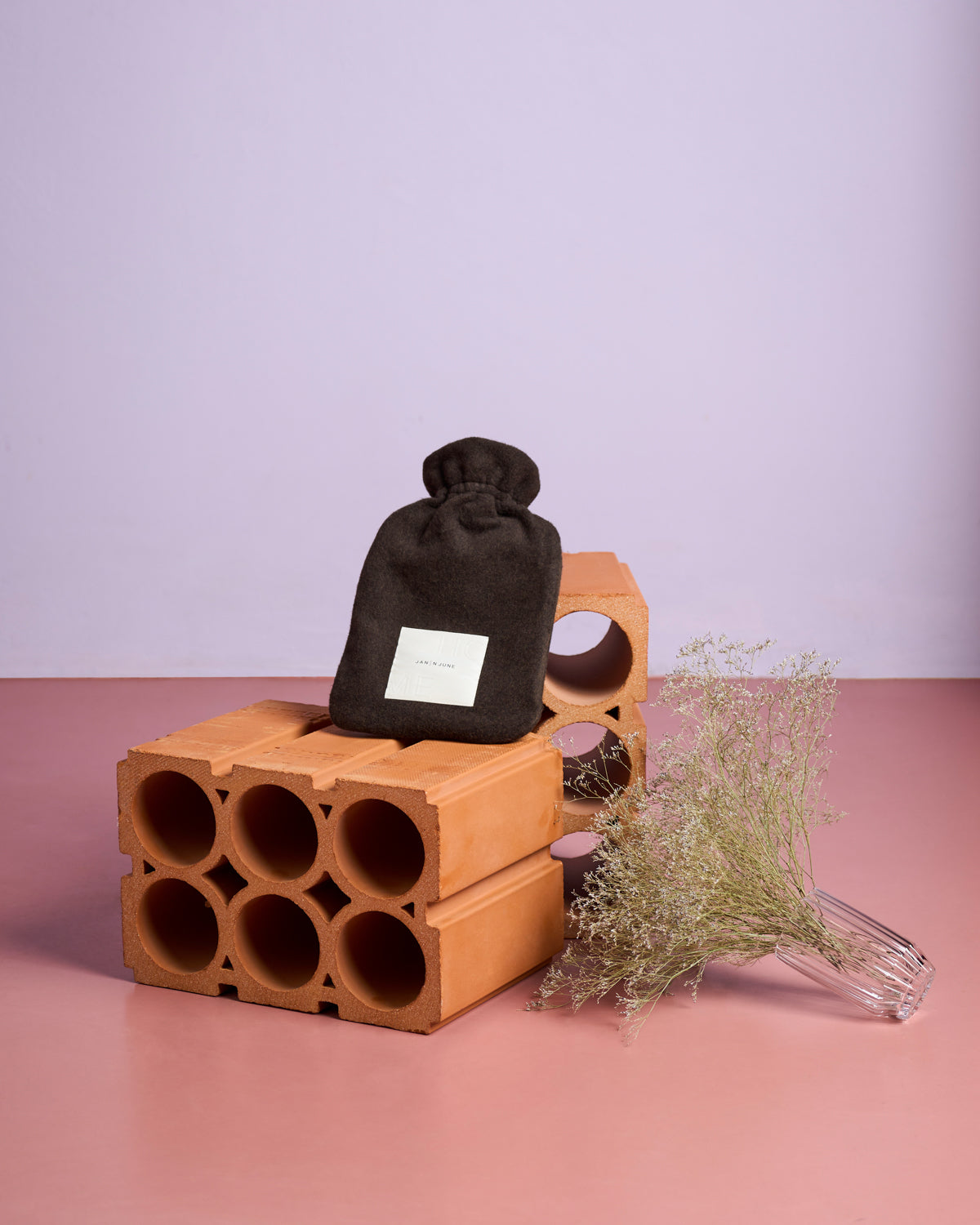 The HOT WATER BOTTLE COVER MOCCA by jan-n-june, crafted from sustainable materials, is displayed on stacked terracotta bricks beside a glass vase of dried flowers, set against a pink floor and pale purple wall backdrop.