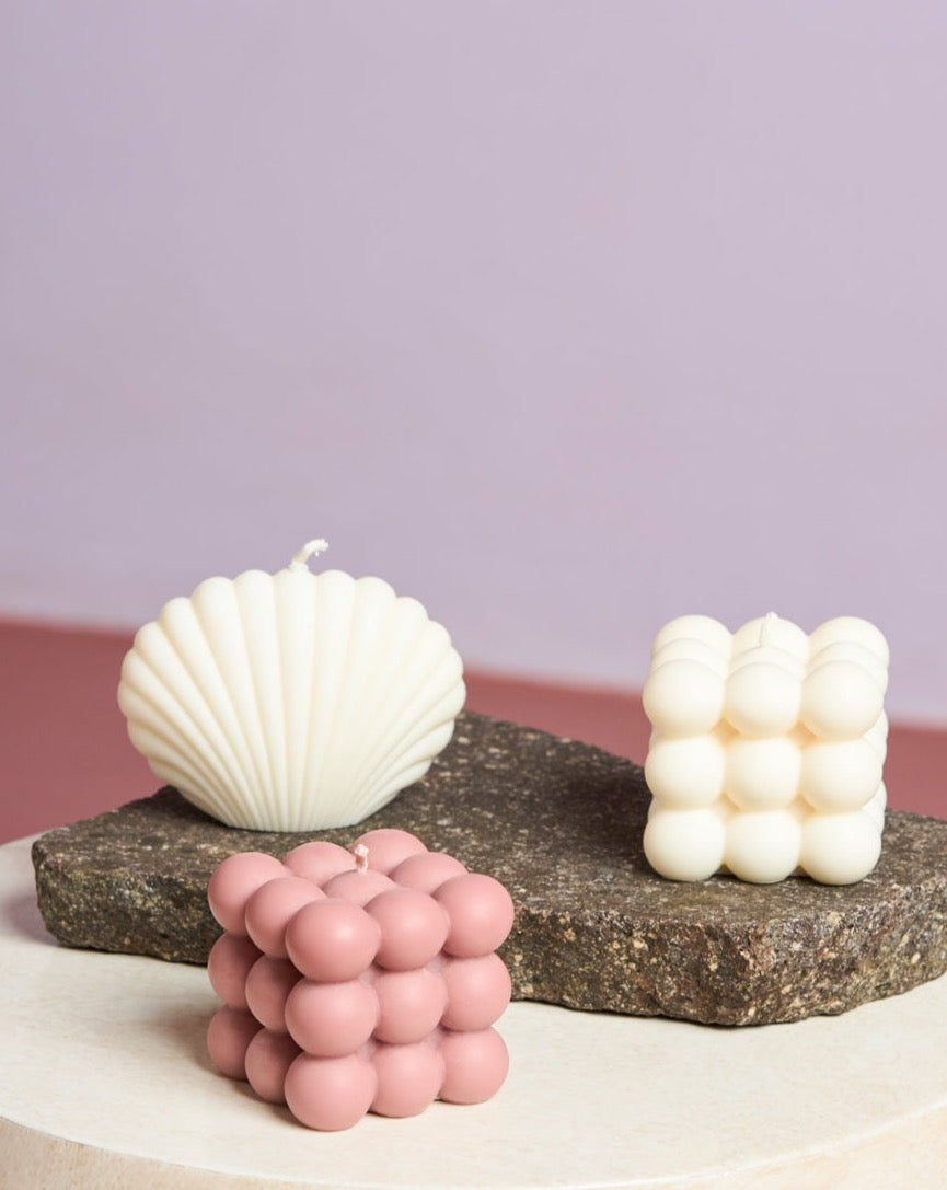 Three decorative candles sit on two stone platforms: a shell-shaped SHELL CANDLE IVORY by jan-n-june, a white bubble cube, and a pink bubble cube—all vegan and eco-friendly—set against a pastel pink and purple background.