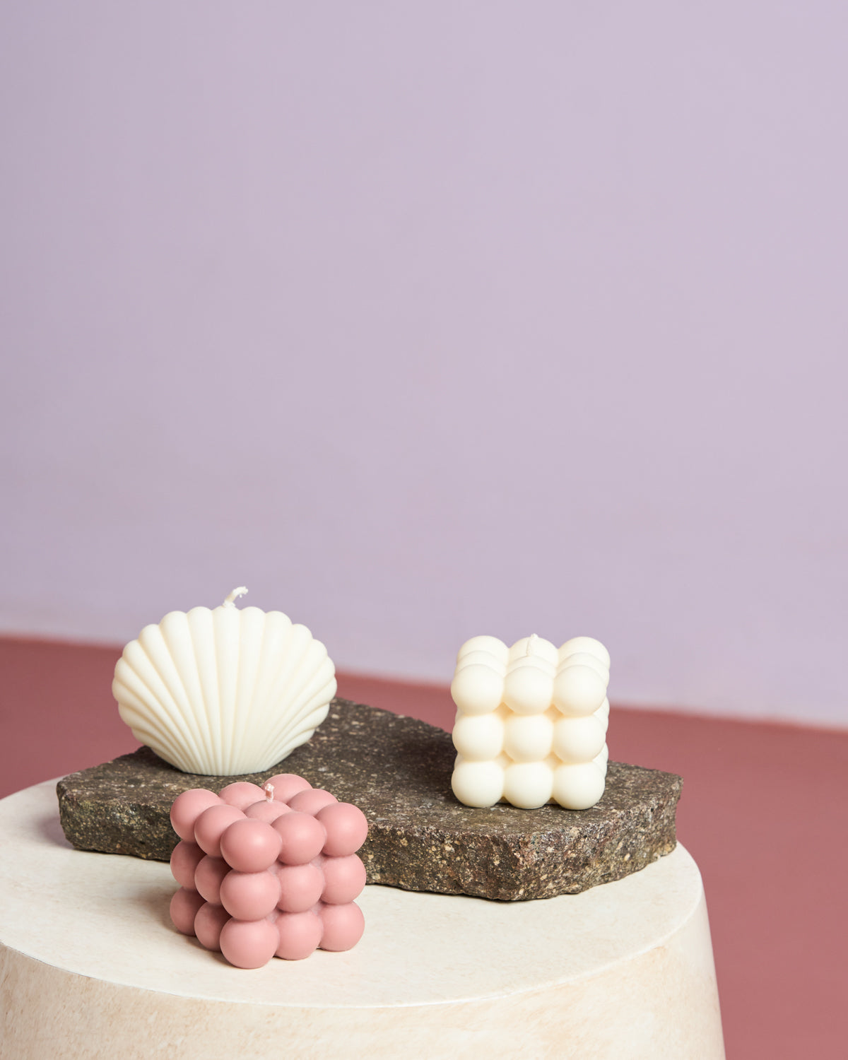 Three handmade candles rest on textured surfaces: a white seashell-shaped candle, jan-n-june’s white BUBBLE TROUBLE CANDLE, and the pink BUBBLE TROUBLE CANDLE ROSE, all set against a soft purple and muted pink background.
