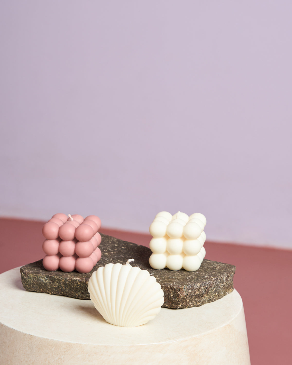 Two cube candles (pink and white) and the jan-n-june SHELL CANDLE IVORY are arranged on textured stone slabs against a pink and purple backdrop. These vegan, eco-friendly accents add charm to any space.