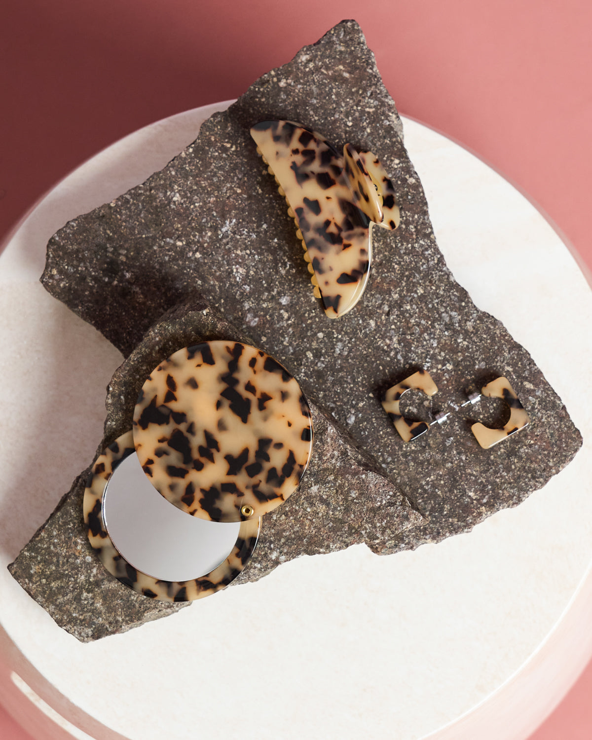 The jan-n-june CIRCLE MIRROR BLONDE TORTOISE, a compact tortoiseshell-patterned accessory, is shown with geometric earrings on a stone slab, set against a pink and white backdrop.