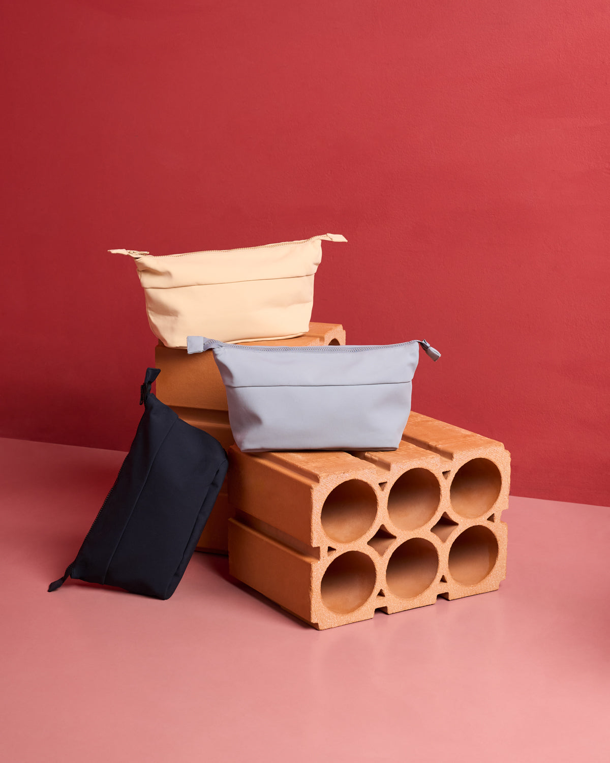 Three rectangular jan-n-june COSMETIC BAGS in cream, light gray, and black with neoprene texture are arranged on and around two stacked hollow bricks set against a pink and red background.