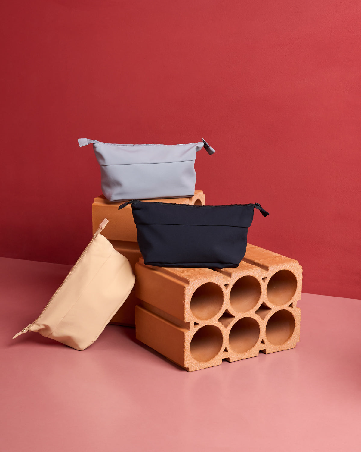 Jan-n-june COSMETIC BAG, in light blue, beige, and black recycled polyamide with a neoprene finish, is shown on stacked clay bricks against a pink and red backdrop.