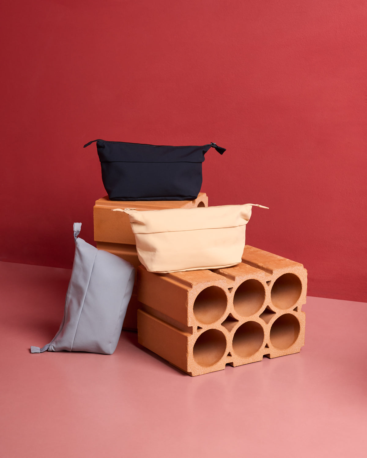 Three jan-n-june COSMETIC BAGs in black, beige, and light gray with a soft neoprene texture are arranged on and around stacked hollow bricks against a pink and red backdrop.