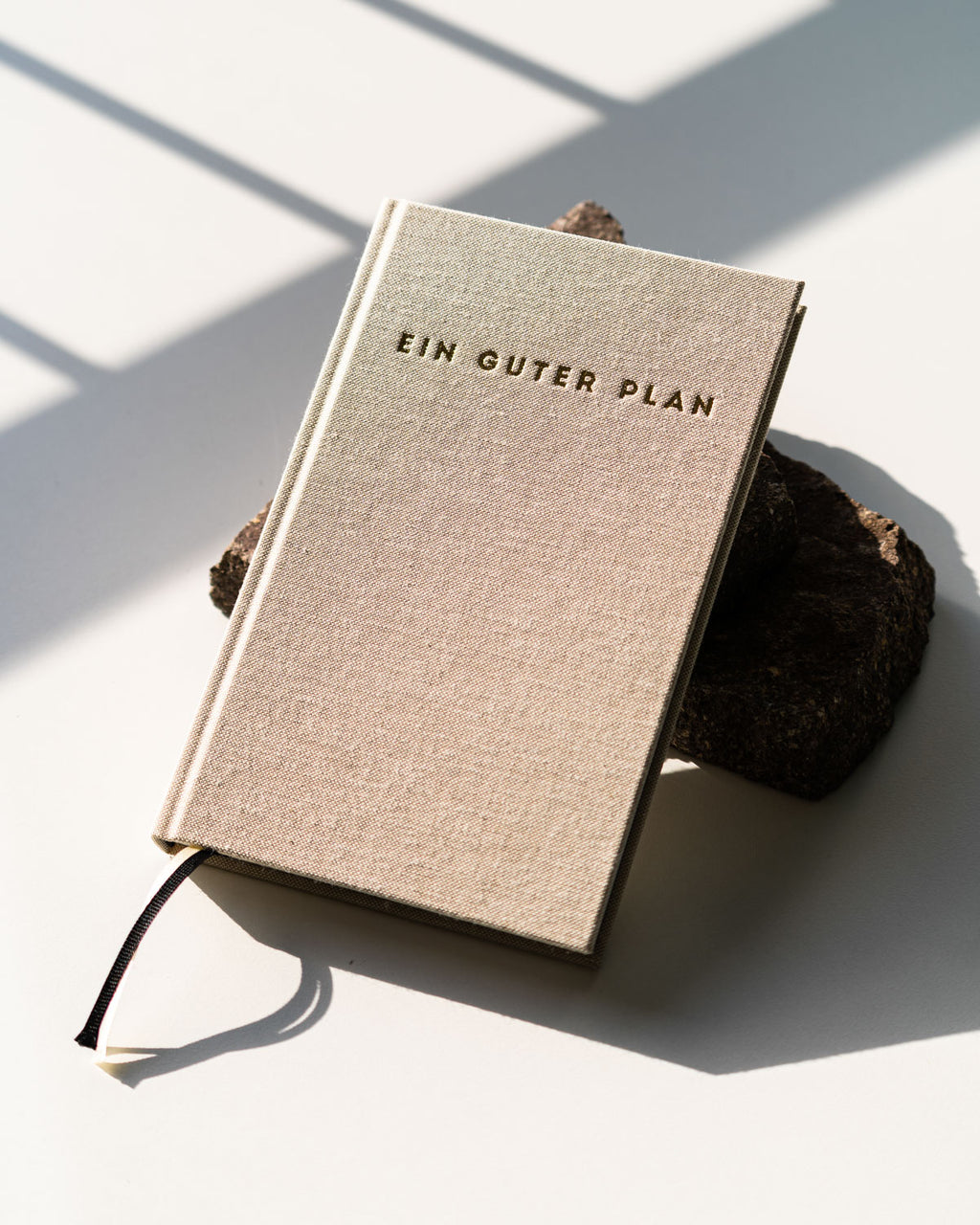 The jan-n-june KALENDER EIN GUTER PLAN NATUR UNDATED, a beige hardcover holistic diary for mindfulness and self-love, sits on two rough stones with sunlight and shadows on a white surface.