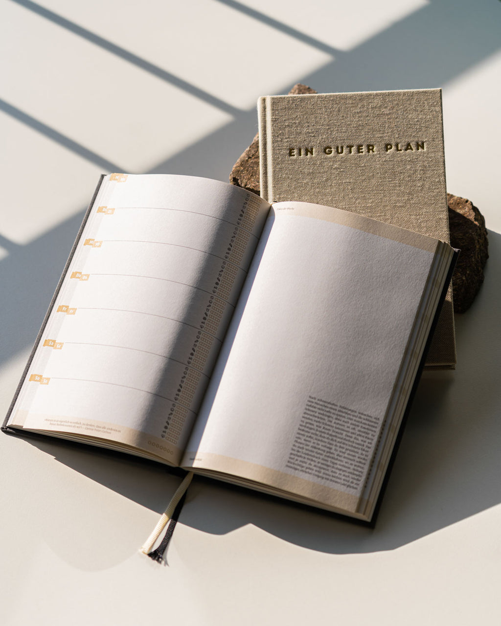 Two beige KALENDER EIN GUTER PLAN NATUR UNDATED planners by jan-n-june sit in sunlight—one closed with embossed cover, the other open to clean, undated pages—ideal for mindful, holistic organization.