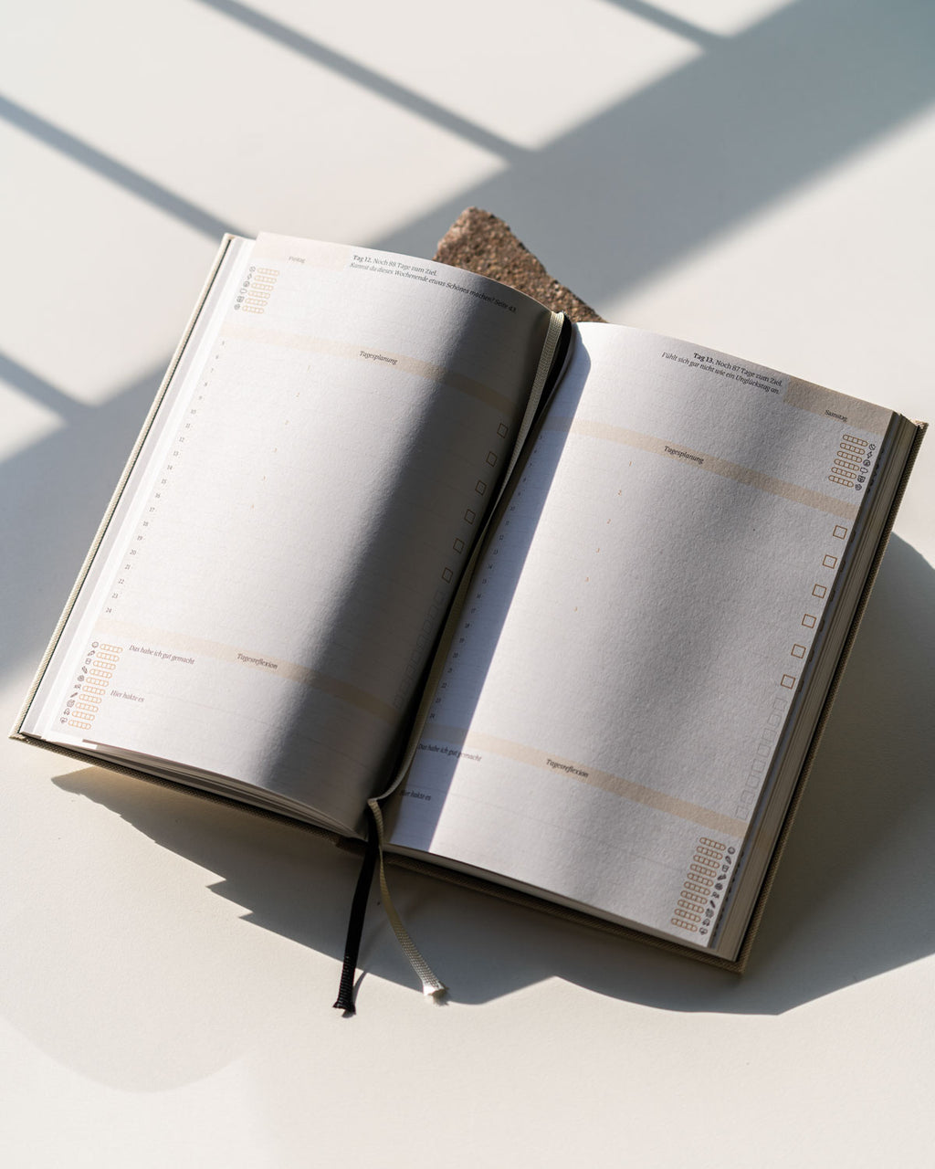 The PLANER EIN GUTES PROJEKT NATUR by jan-n-june, an open eco-friendly journal with blank pages, lies on a white surface dappled in sunlight and windowpane shadows—a brown item keeps this sustainably made German planner open.