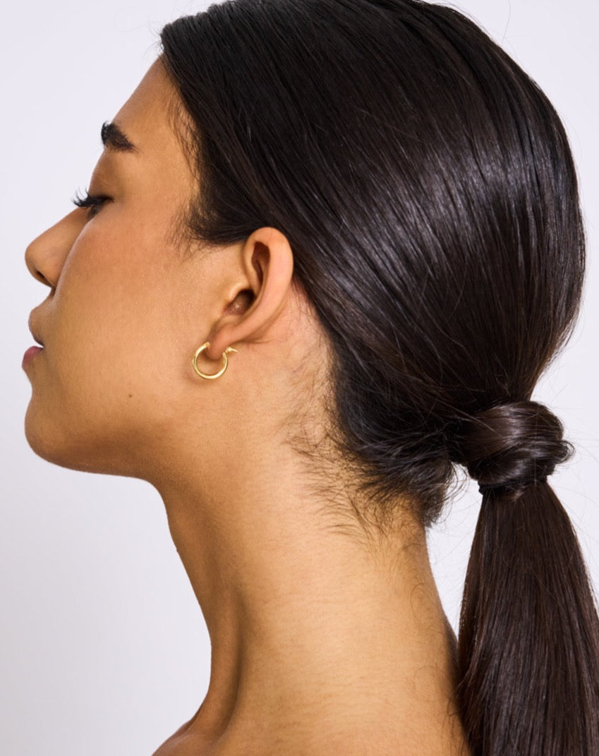 A woman with straight dark hair in a low ponytail wears jan-n-june’s DEAR DARLING BERLIN ESSENTIAL HOOP EARRINGS SMALL GOLD, crafted from recycled 925 Sterling Silver, shown in profile against a plain light background.