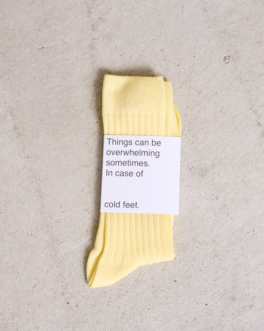 A pair of folded SOCKS FADED LEMON by jan-n-june in yellow rests on a light textured surface, wrapped with a label that reads: Things can be overwhelming sometimes. In case of cold feet.