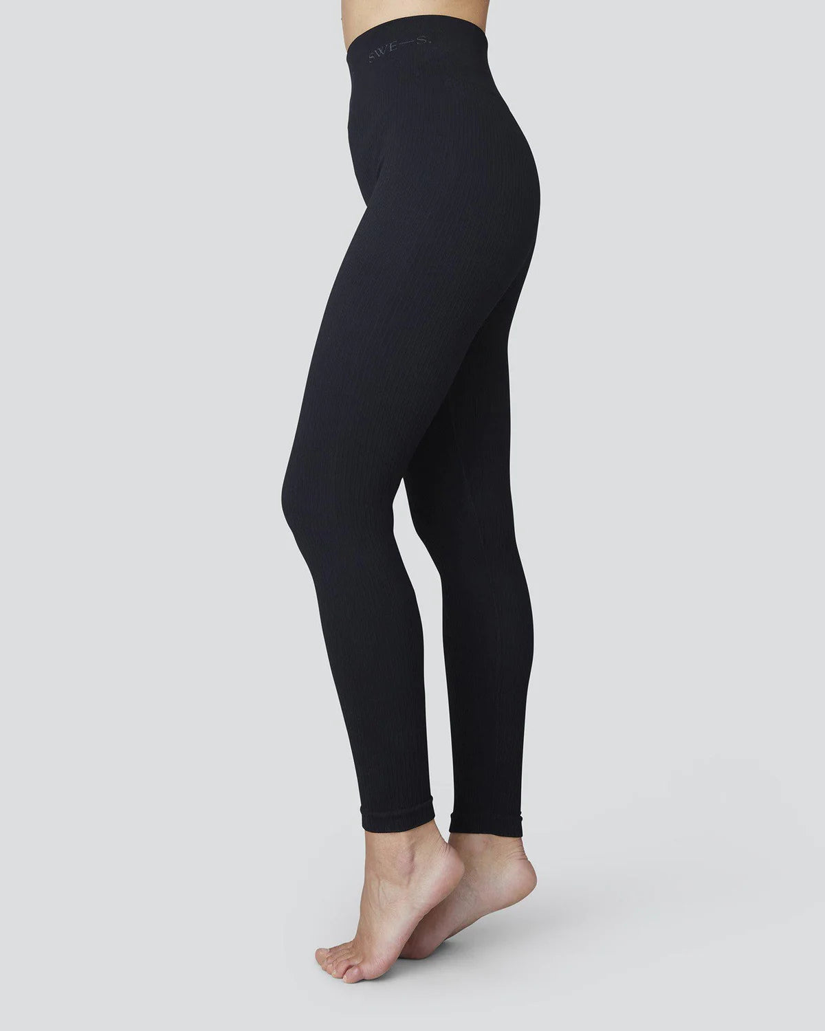 A person stands barefoot on tiptoe against a plain light gray background, wearing jan-n-june SWEDISH STOCKINGS TYRA RIB LEGGINGS BLACK—seamless, high-waisted leggings with a broad monogrammed waistband made from recycled yarns.