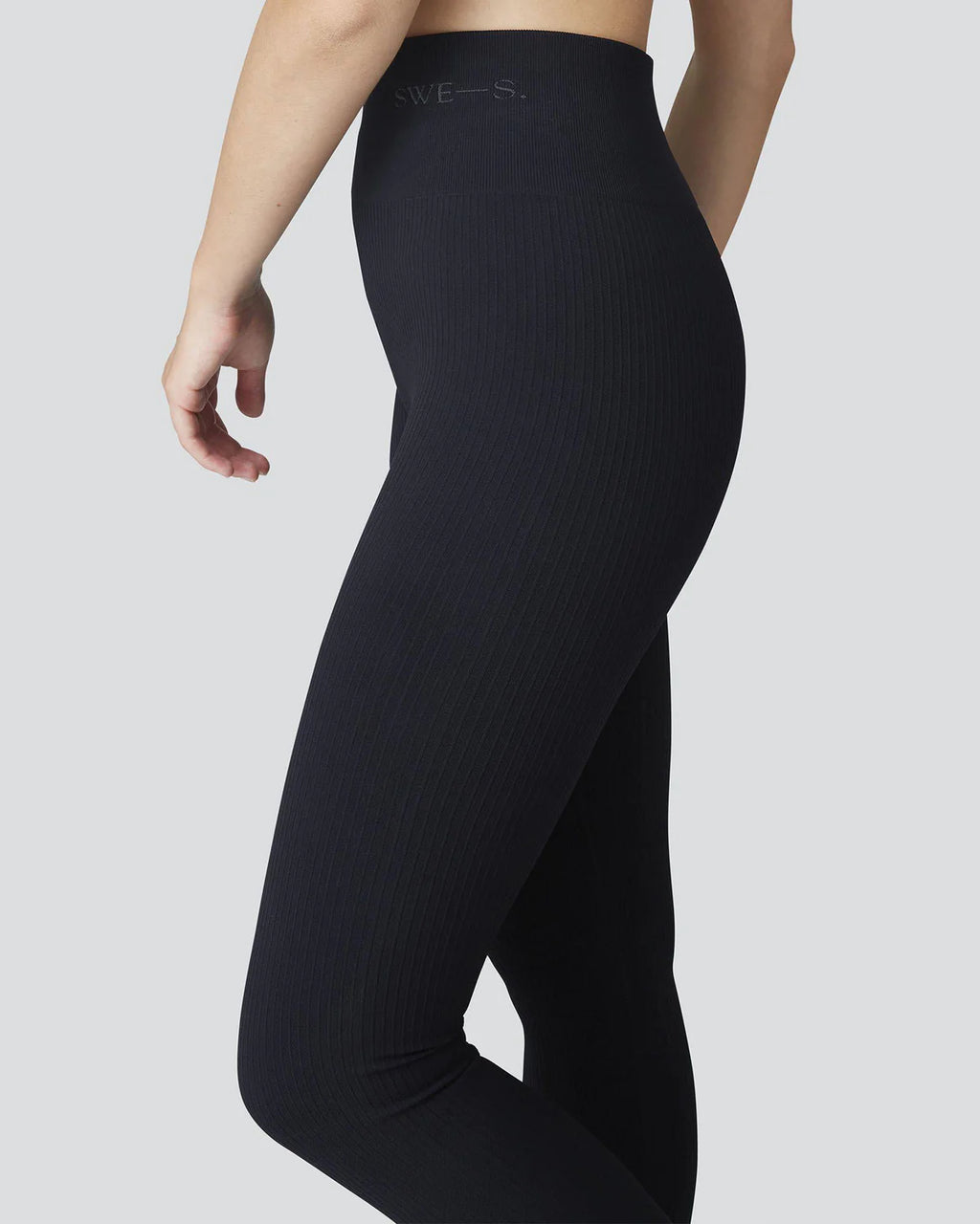 A model, shown from mid-torso to ankles and standing sideways against a plain light gray background, wears jan-n-junes SWEDISH STOCKINGS TYRA RIB LEGGINGS BLACK featuring a high waist, seamless ribbed design, and broad monogrammed waistband.