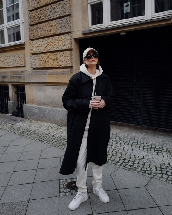 On a city sidewalk, a person wears the jan-n-june COAT TAGUS BLACK with an oversized cut over a white hoodie and sweatpants, white sneakers, a white cap, sunglasses, and holds a coffee cup.