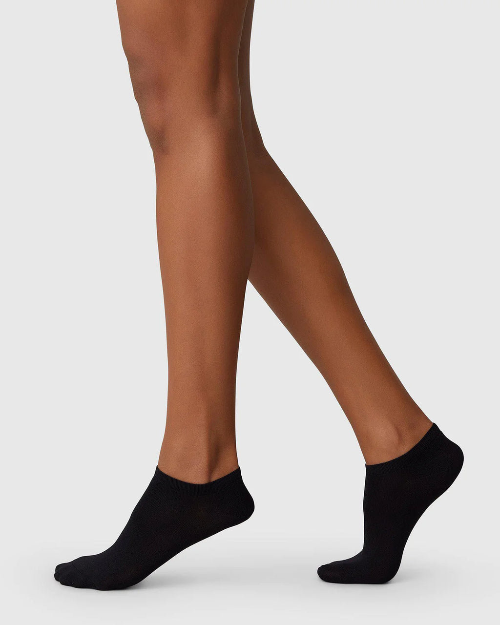 A person wears jan-n-june SWEDISH STOCKINGS SOCKS SARA BLACK, made from recycled yarn, standing on tiptoe with one leg slightly bent, against a plain light gray background.