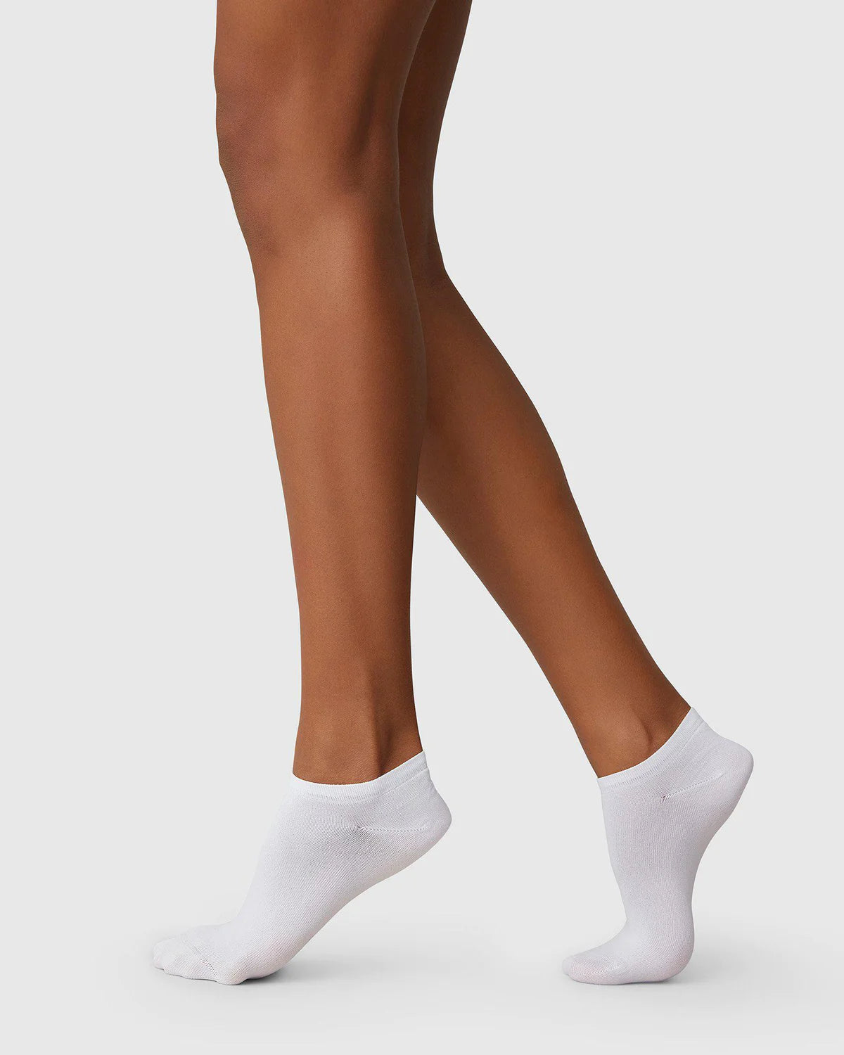 A person with medium to dark skin tone wears jan-n-june SWEDISH STOCKINGS SOCKS SARA WHITE, made from recycled yarn, standing with one foot slightly in front of the other against a plain light gray background.