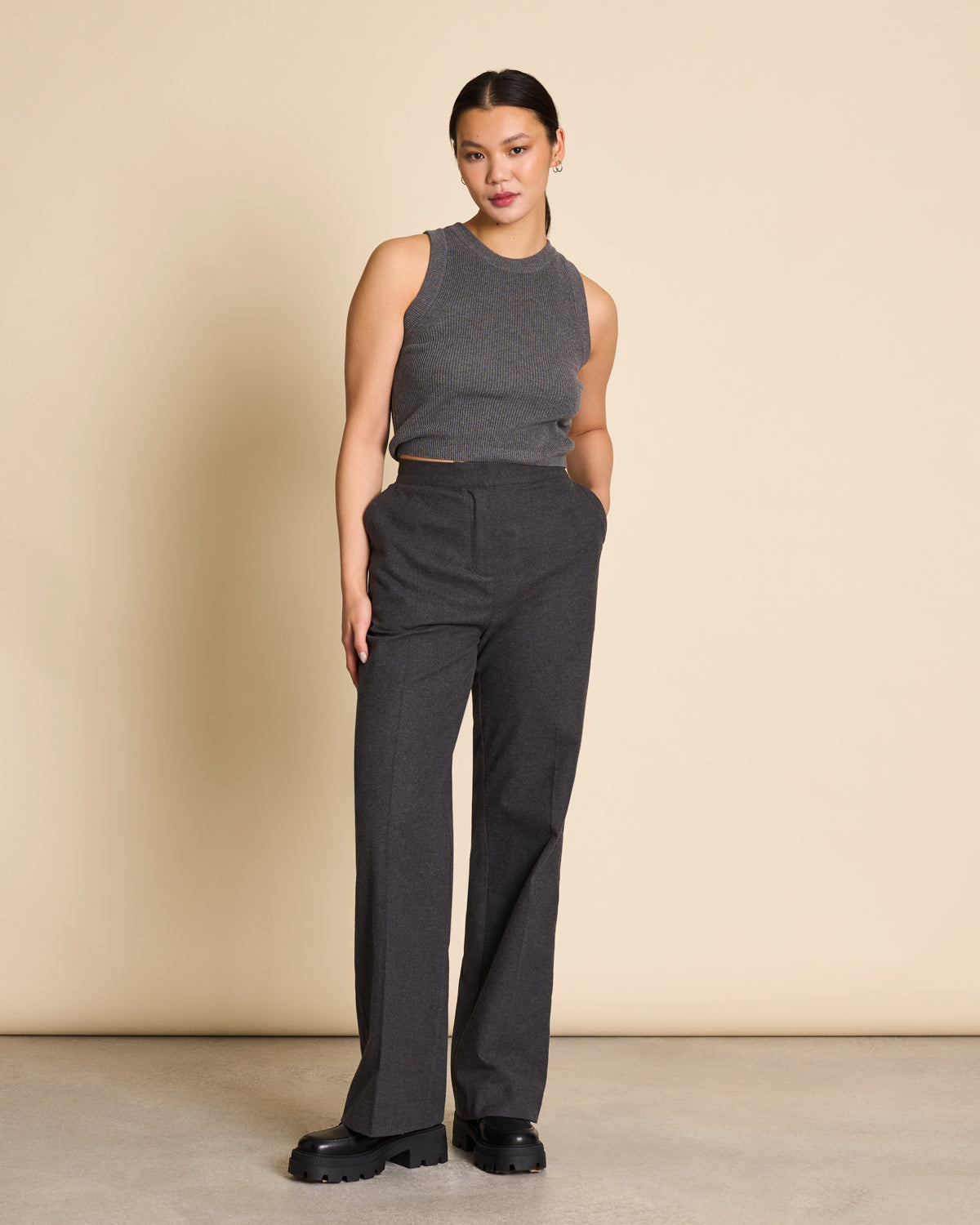 A woman stands against a beige background, confidently looking at the camera in black chunky shoes. She wears jan-n-june’s KNIT TOP GARDA DARK GREY GOTS paired with high-waisted dark gray trousers, her left hand casually in her pocket.