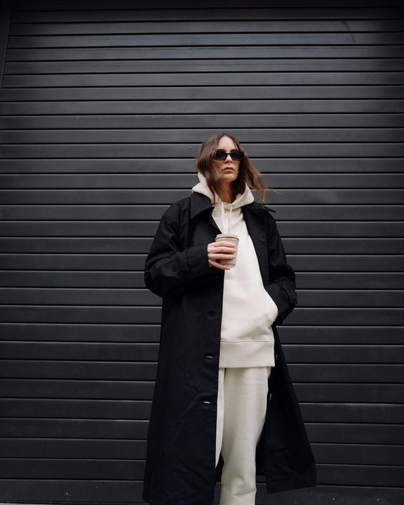 Wearing the jan-n-june COAT TAGUS BLACK, a woman stands before a dark garage door, layering the long, oversized coat over a cream hoodie and sweatpants, accessorized with sunglasses and holding a coffee cup.