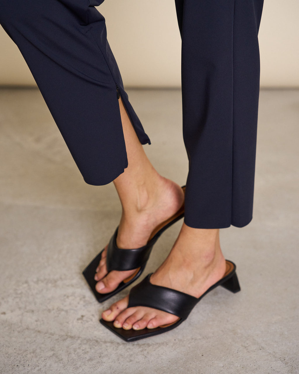 A person wears jan-n-june PANTS STRAIGHT BLACK—high waisted, straight-leg, sustainable fabric with hidden hem zippers—and black open-toe low-heeled sandals, standing on a light-colored concrete floor.