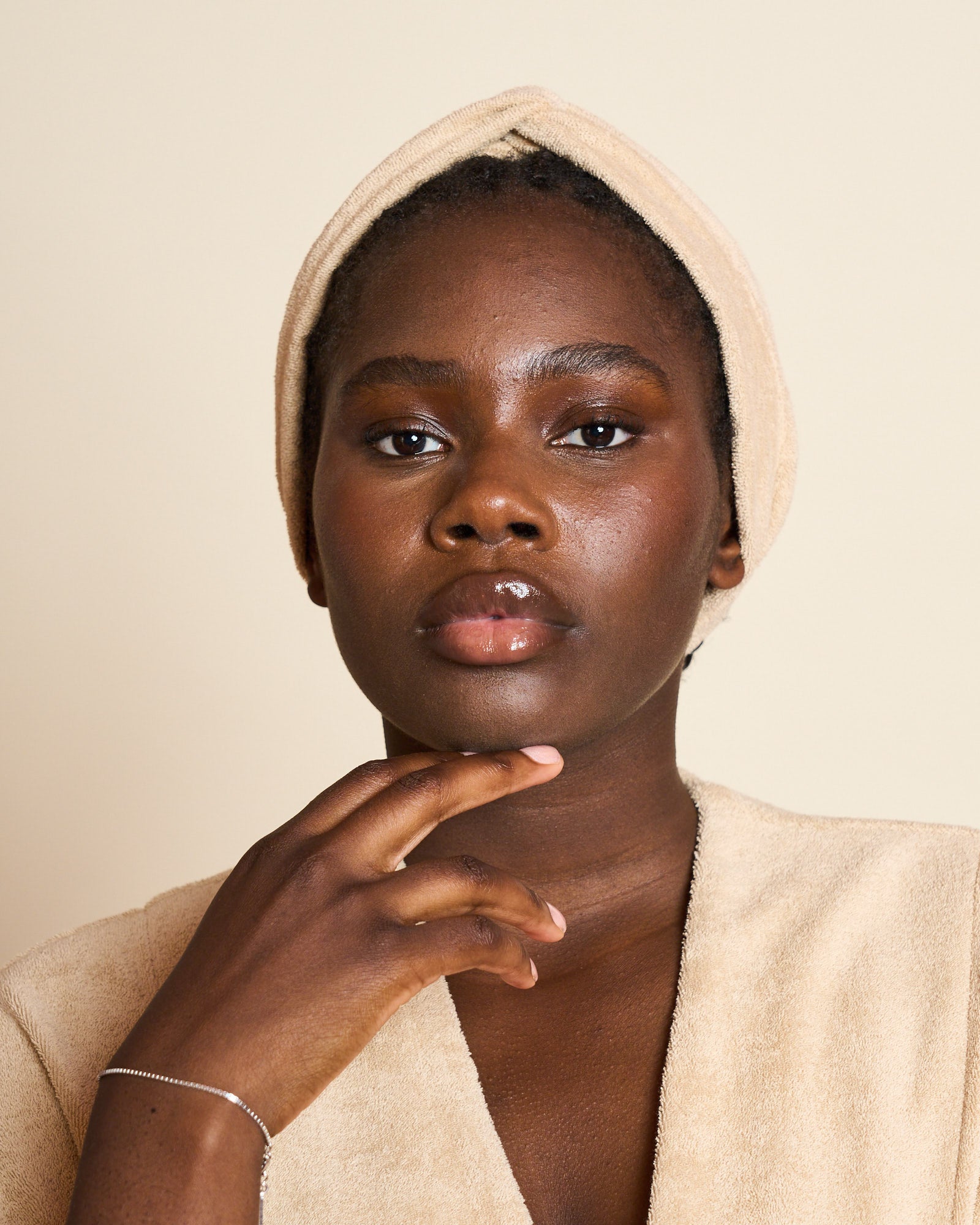 A woman with medium-dark skin, wearing a beige towel around her head and shoulders, gazes calmly at the camera, resting her hand gently on her chin against a neutral background.