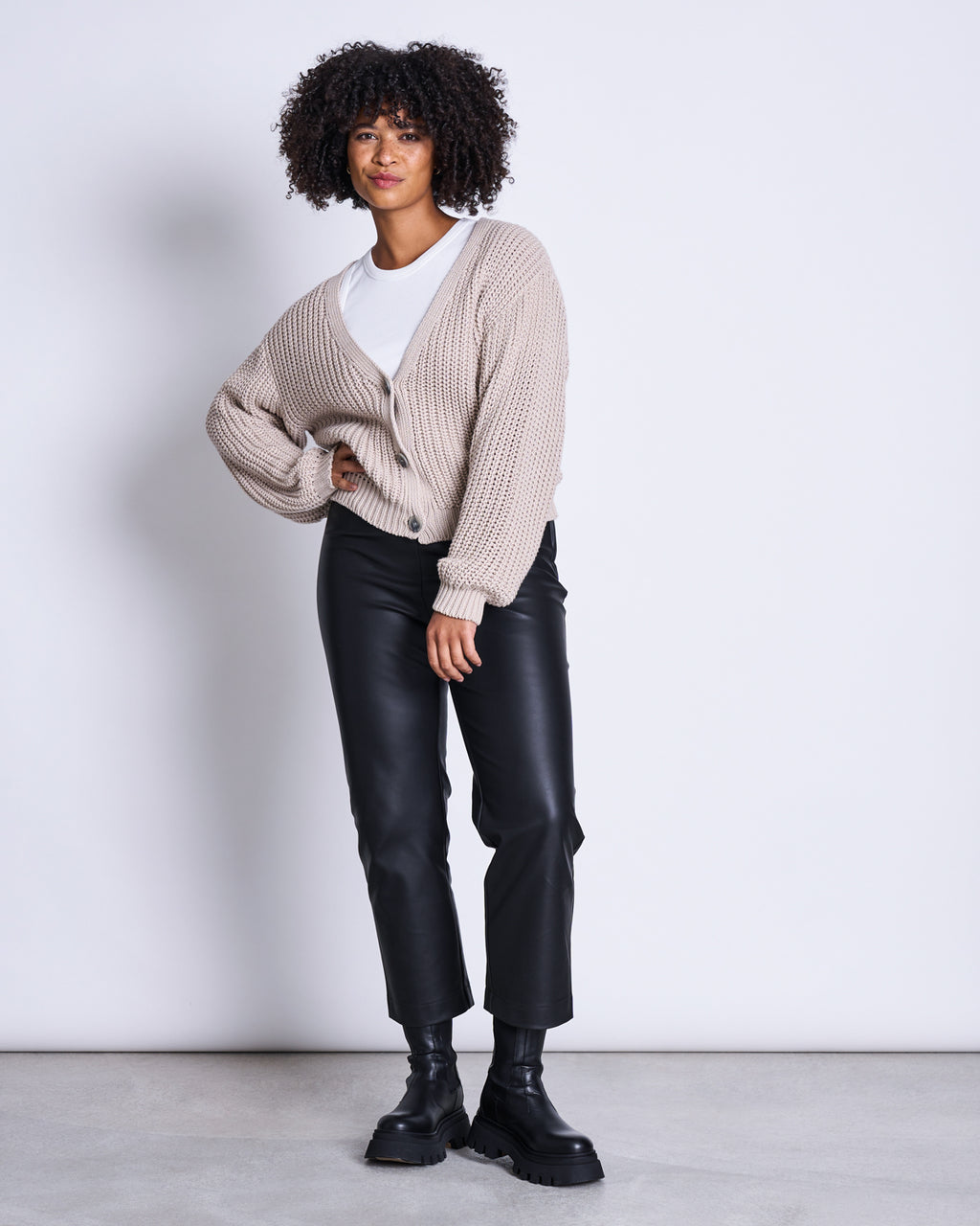 A person with curly hair wears the jan-n-june CHUNKY CARDIGAN LENA IVORY GOTS over a white shirt, black leather pants, and black chunky boots, standing in front of a plain light background.