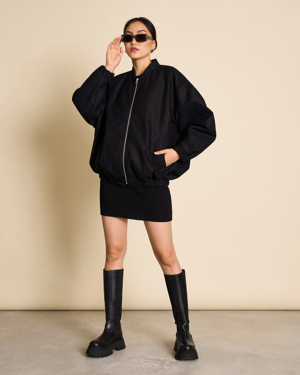 A person wearing sunglasses, a loose black zip-up jacket, the jan-n-june SKIRT LOKKA BLACK GOTS with an elasticated waistband, and knee-high black chunky boots stands confidently against a beige background.