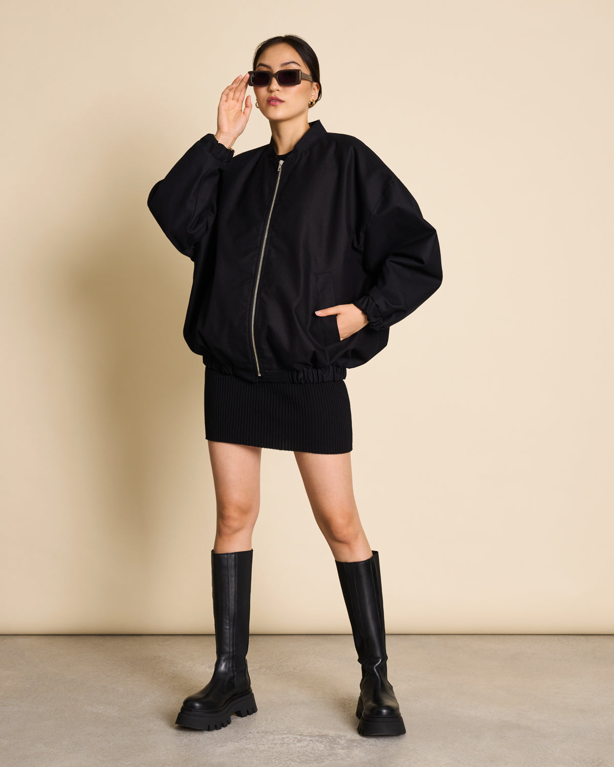 A person wearing sunglasses, a loose black zip-up jacket, the jan-n-june SKIRT LOKKA BLACK GOTS with an elasticated waistband, and knee-high black chunky boots stands confidently against a beige background.