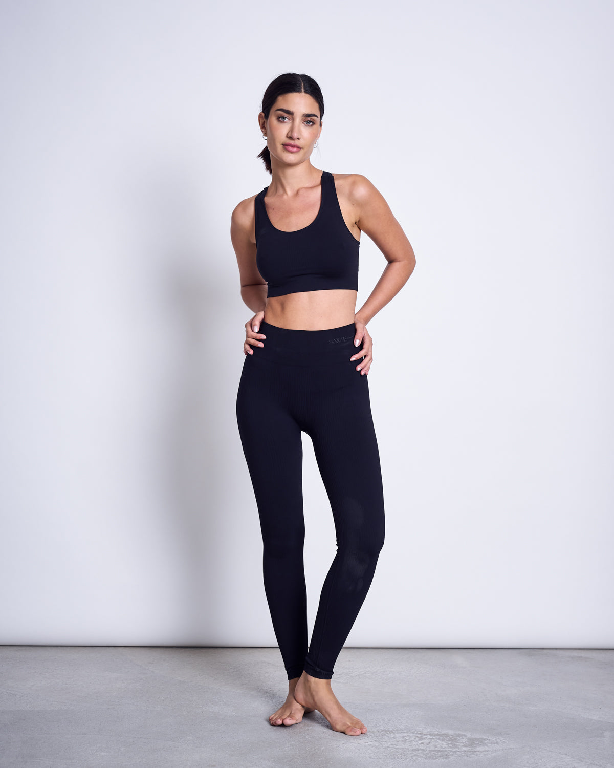 A woman stands barefoot on a gray floor against a white wall, wearing jan-n-junes SWEDISH STOCKINGS TYRA RIB LEGGINGS BLACK and a black sports bra, her hair tied back with one hand on her hip.