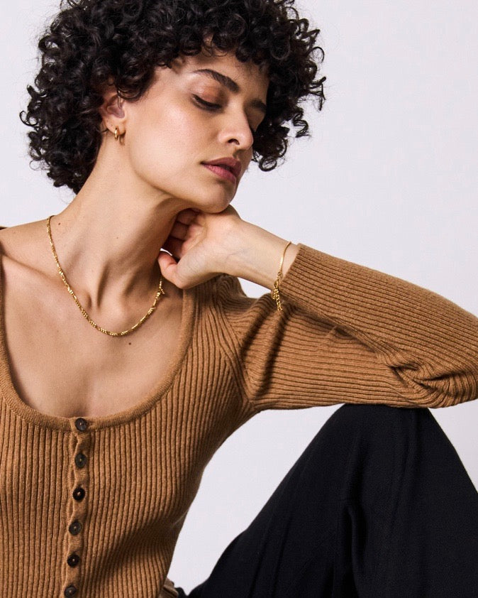 A woman with short curly hair wears a ribbed tan button-up top, black pants, gold jewelry—including the jan-n-june DEAR DARLING BERLIN FIGARO CHAIN NECKLACE crafted from recycled 925 sterling silver—posed against a plain background.