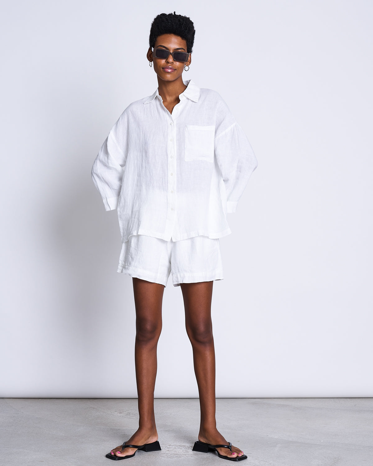 A person stands against a plain background wearing jan-n-june’s LINEN SHIRT MONS WHITE, paired with sunglasses, white shorts, hoop earrings, and black sandals. Hands in pockets, they have a relaxed expression.
