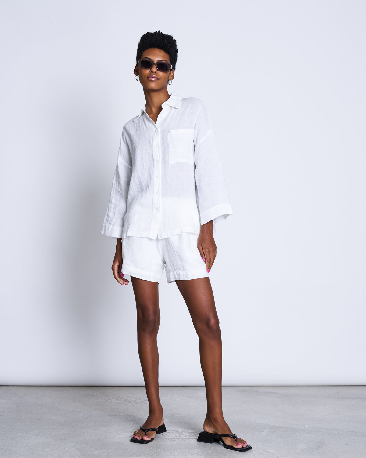 A person in sunglasses and black sandals confidently poses against a white background, wearing the jan-n-june LINEN SHIRT MONS WHITE—a loose European linen button-up with a classic collar—and matching white shorts.