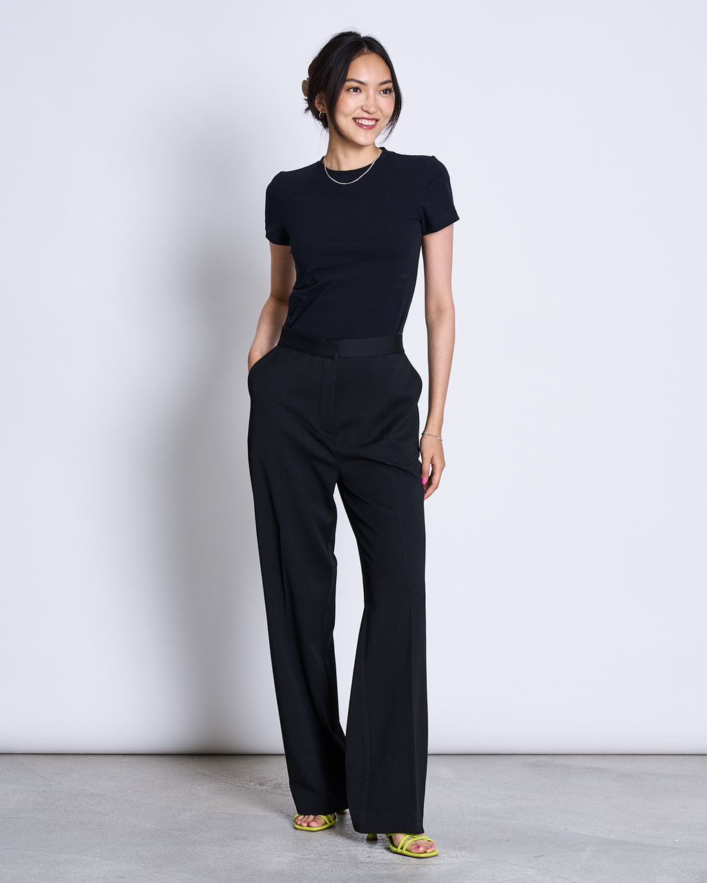 A woman stands against a light background, smiling with hands in her pockets, wearing the jan-n-june T-SHIRT PALOOMA BLACK GOTS and black wide-leg trousers, paired with yellow open-toe heels.