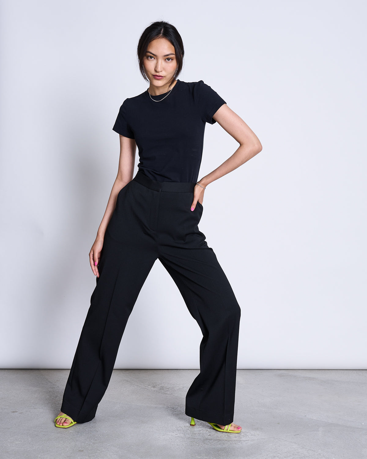 A woman stands confidently on a concrete floor, wearing the jan-n-june T-SHIRT PALOOMA BLACK GOTS and black wide-leg pants, paired with yellow heeled sandals, posing against a plain light gray background.