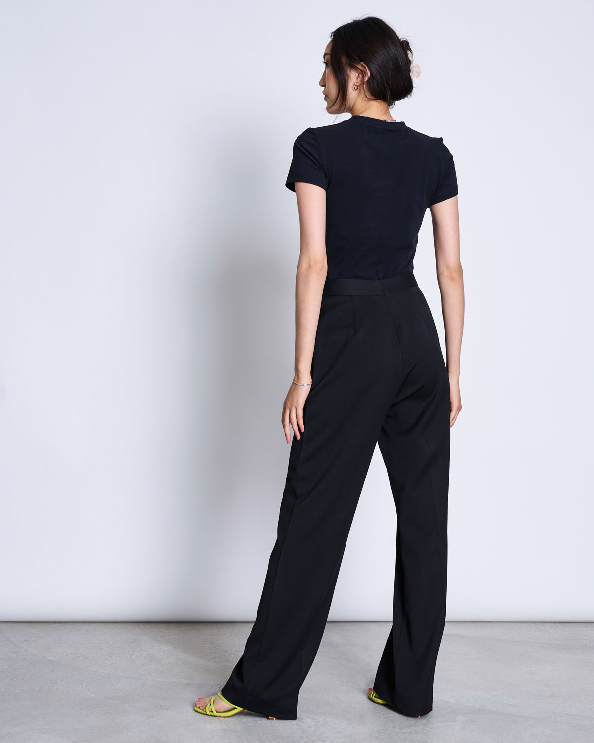 A woman faces away from the camera, wearing the jan-n-june T-SHIRT PALOOMA BLACK GOTS with high-waisted black trousers and yellow-heeled sandals, set against a plain light background.